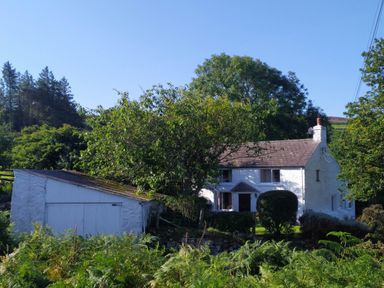A white house with a chimney and an adjacent white garage surrounded by trees and greenery at Penllain in Newport Pembrokeshire