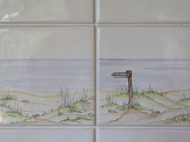 A painted beach scene with sand dunes and grass with a wooden signpost reading Penllain on ceramic tiles at Penllain in Newport Pembrokeshire