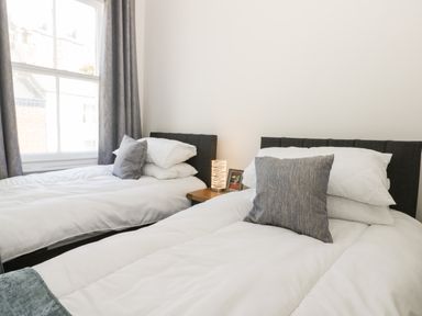 A bedroom with two single beds white bedding gray pillows a window with gray curtains and a wooden nightstand with a lamp and picture frame at Rydal Cottage in Scarborough
