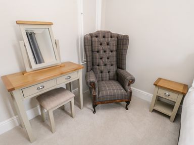 A room corner with a tiled armchair a wooden dressing table with mirror and stool and a small wooden bedside table at Rydal Cottage in Scarborough