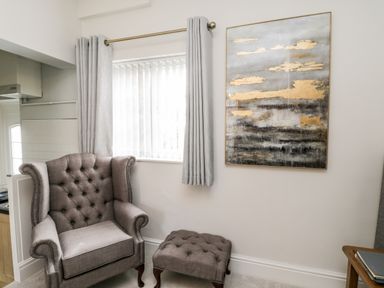 A grey upholstered armchair with a matching footstool near a window with grey curtains and an abstract painting on the wall at Rydal Cottage in Scarborough