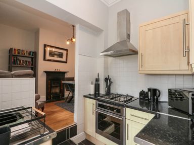 A kitchen with a gas stove oven and black countertop adjoining a room with a bookshelf and dining table at Rydal Cottage in Scarborough