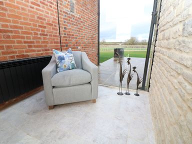 A gray armchair with a blue floral pillow on a tiled floor between a brick wall and a stone wall with three metal bird sculptures near a glass door
