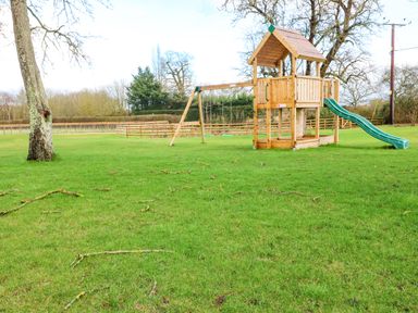 A garden with a wooden climbing frame slide and swings on grass with trees and fencing at Morgans Farmhouse in Bampton Oxfordshire