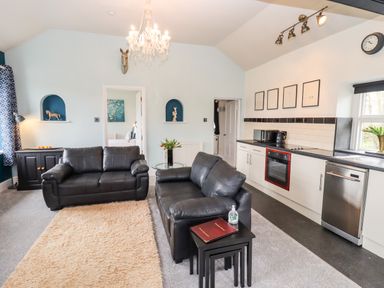 A living room with black leather sofas a kitchen area with oven and dishwasher and a chandelier at The Stable in Lydney