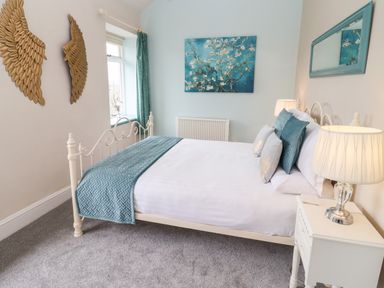 A bedroom with a white bed frame blue bedding and pillows a nightstand with a lamp wall decorations including a gold wing sculpture and a framed painting at The Stable in Lydney