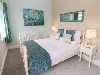 A bedroom with a bed with blue and white bedding two bedside tables with lamps a radiator a window with blue curtains and a painting above the radiator at The Stable in Lydney
