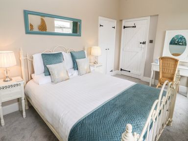 A bedroom with a bed with blue and white bedding two bedside tables with lamps a blue framed mirror a wicker chair and a white vanity at The Stable in Lydney