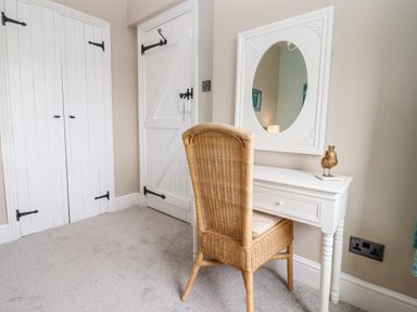 A room with a white dressing table and oval mirror with a wicker chair and white doors at The Stable in Lydney