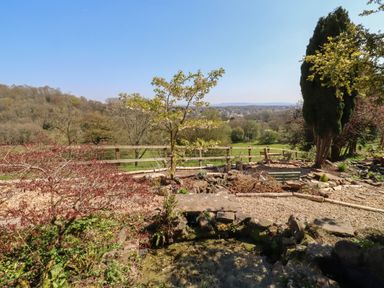 A garden area with trees shrubs a pond and seating with a view of fields and distant town at The Stable in Lydney