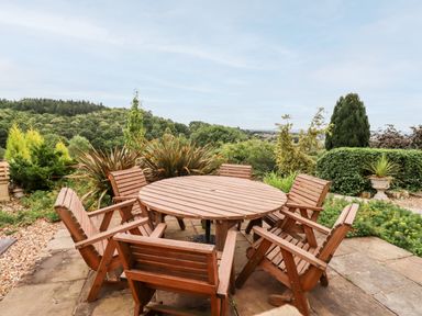 An outdoor patio with a round wooden table and six wooden chairs surrounded by greenery at The Stable in Lydney