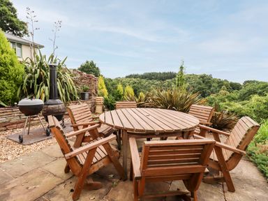An outdoor patio with a round wooden table and six wooden chairs surrounded by plants and trees