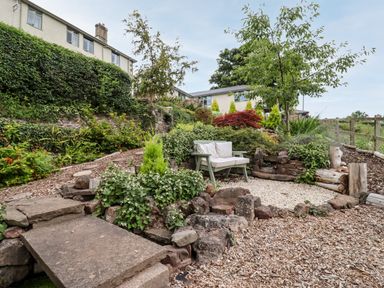 A garden area with a green bench and white cushions surrounded by plants and rocks at The Stable in Lydney