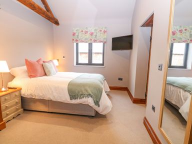 A bedroom with a double bed bedside tables lamps a window with floral curtains a wall mounted TV and a large mirror at Shorthorn Barn in Attlebridge near Taverham