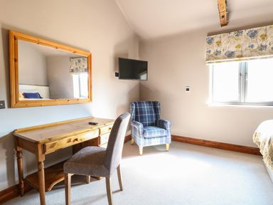 A bedroom with a wooden desk and chair a wall mirror a blue plaid armchair and a window with floral blinds at Shorthorn Barn in Attlebridge near Taverham