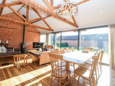 An open living and dining room with wooden beams a brick wall a dining table with chairs sofas a TV and large glass sliding doors at Shorthorn Barn in Attlebridge near Taverham