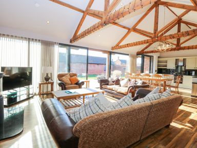 An open living room and kitchen with wooden beams on the ceiling large windows sofas armchair coffee table dining table and chairs a television and kitchen cabinets at Shorthorn Barn Attlebridge near Taverham