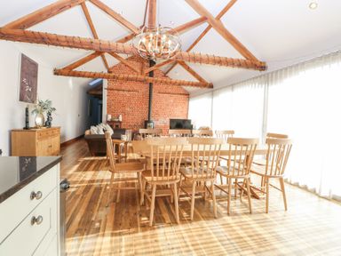 A dining area with a wooden table and chairs beneath exposed wooden beams and a brick wall with a wood stove at Shorthorn Barn in Attlebridge near Taverham