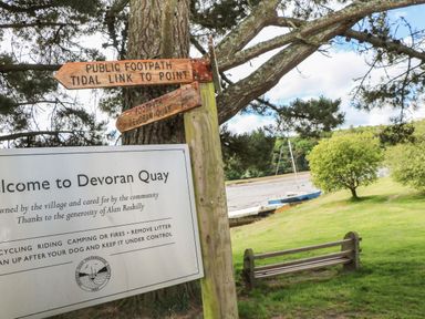 A wooden signpost and a welcome sign near a grassy area with a bench and a boat by the water at Armando in Devoran Cornwall