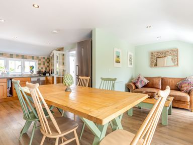 A dining area with a wooden table and chairs next to a kitchen with wooden cabinets and a stainless steel refrigerator and a living area with a brown sofa and coffee table at Armando in Devoran Cornwall