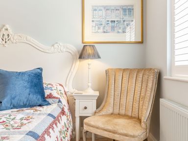 A bedroom corner with a white ornate headboard a colorful quilt and a blue pillow a white nightstand with a lamp and a beige upholstered chair at Armando in Devoran Cornwall