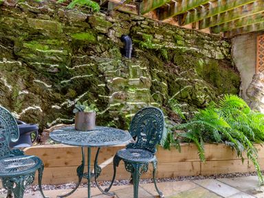 An outdoor seating area with a metal table and two chairs next to a stone wall with moss and plants at Armando in Devoran Cornwall
