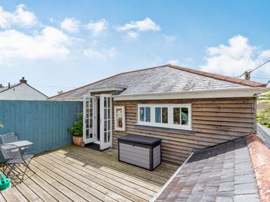An outdoor wooden deck with a small table and two chairs next to a wooden fence and a building with French doors and windows at Armando in Devoran Cornwall