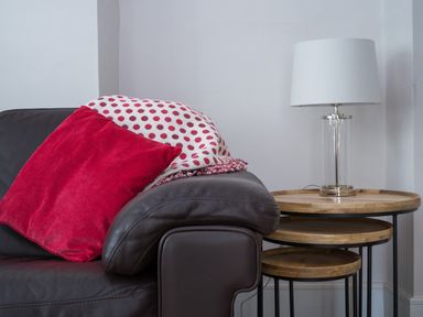 A dark brown leather sofa with red pillow and white and red polka dot blanket next to nested wooden side tables with a lamp