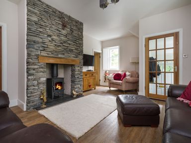A living room with a stone fireplace a wood stove leather sofas a striped armchair a wooden cabinet and a glass paneled door at Fellside Lodge in Bowness-On-Windermere