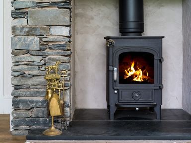 A wood burning stove with fire inside next to a stone wall and brass fireplace tools at Fellside Lodge in Bowness-On-Windermere