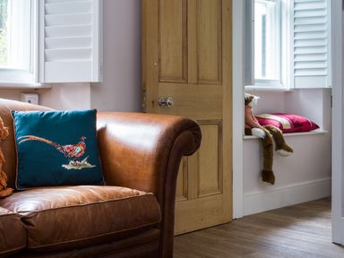 A leather sofa with a pillow with a bird design and a wooden door next to a window seat with a red pillow and a stuffed animal at Fellside Lodge in Bowness-On-Windermere