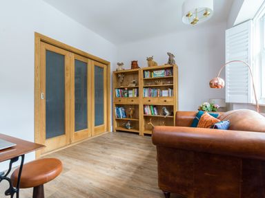 A room with a leather sofa, wooden bookshelves holding books and stuffed animals, wooden sliding doors, a floor lamp, and a small table with a stool at Fellside Lodge in Bowness-On-Windermere