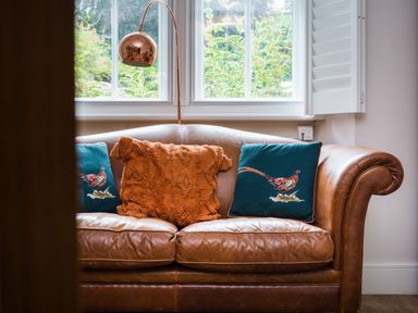 A brown leather sofa with decorative cushions and a brass floor lamp next to a window at Fellside Lodge in Bowness-On-Windermere