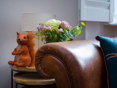A brown leather sofa with a dark blue cushion next to a wooden side table holding a lamp a bouquet of flowers and a stuffed squirrel at Fellside Lodge in Bowness-On-Windermere