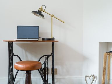 A small workspace with a wooden desk, a laptop, a desk lamp, and a brown round stool at Fellside Lodge in Bowness-On-Windermere