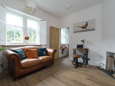 A living room with a brown leather sofa pillows a wooden door a small wooden desk with a laptop stool and a painting of a bird on the wall at Fellside Lodge in Bowness-On-Windermere