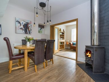 A dining area with a wooden table and leather chairs next to a wood-burning stove and an adjoining room with a bookshelf and armchair at Fellside Lodge in Bowness-On-Windermere
