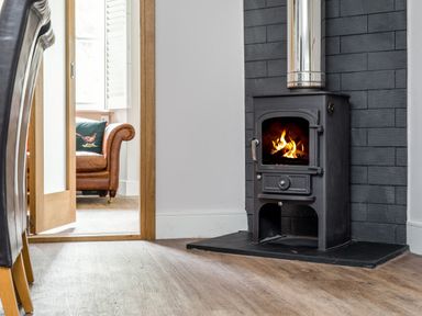A room with a wood burning stove next to a doorway showing a leather armchair at Fellside Lodge in Bowness-On-Windermere
