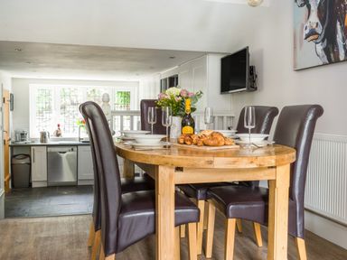 A dining area with four dark leather chairs around a wooden table set with plates glasses and pastries in a kitchen at Fellside Lodge Bowness-On-Windermere