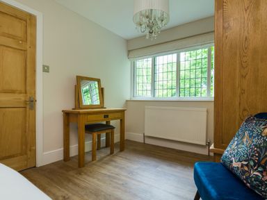 A bedroom with a wooden dressing table and chair next to a window and a blue chair with a patterned cushion at Fellside Lodge in Bowness-On-Windermere