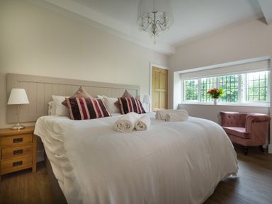 A bedroom with a bed white linens and striped pillows beside a wooden nightstand and a window with a flower vase at Fellside Lodge in Bowness-On-Windermere