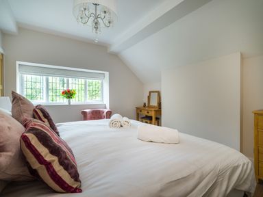 A bedroom with a bed, pillows, folded towels, a dressing table, an armchair, a window, and a chandelier at Fellside Lodge in Bowness-On-Windermere