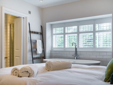 A bedroom with white towels on the bed a freestanding bathtub under a window with shutters and a towel ladder at Fellside Lodge in Bowness-On-Windermere