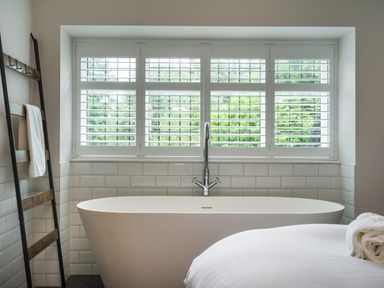 A bathroom with a freestanding bathtub under a window with shutters and a towel on a wooden ladder at Fellside Lodge in Bowness-On-Windermere