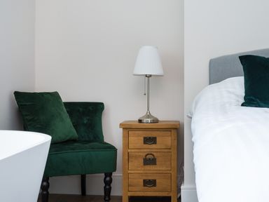 A bedroom corner with a green armchair next to a wooden bedside table with a lamp and part of a bed with white bedding at Fellside Lodge in Bowness-On-Windermere