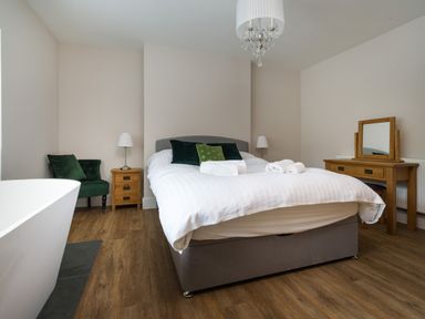 A bedroom with a double bed with pillows and towels bedside tables a green chair a dressing table with a mirror and a chandelier at Fellside Lodge in Bowness-On-Windermere