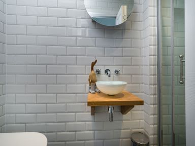 A small bathroom with white subway tiles a round mirror a wooden shelf with a white vessel sink and a wooden duck figurine Fellside Lodge Bowness-On-Windermere