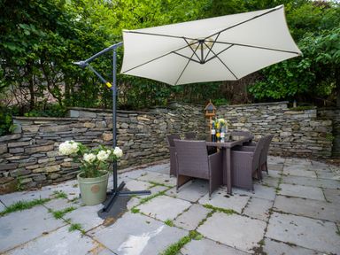 An outdoor patio with a stone wall six chairs around a table and a large white umbrella and a potted plant at Fellside Lodge in Bowness-On-Windermere