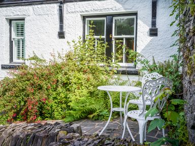 An outdoor seating area with a white metal table and two chairs next to green plants and a white building with black window frames at Fellside Lodge in Bowness-On-Windermere