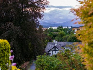 A house surrounded by trees and bushes with mountains and a lake in the background at Fellside Lodge in Bowness-On-Windermere
